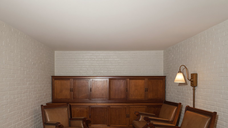 A finished lower-level snug beneath a Federal-era Beacon Hill Boston rowhouse: low flat white ceiling, warm white painted brick foundation walls, warm leather armchairs, dark walnut built-in cabinets, soft brass lamplight