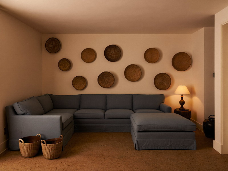A warm lower-level family room beneath an older rowhouse: low flat ceiling with no windows, soft cream walls, deep gray-blue sectional, plain woven baskets with no tags, cork floor tiles, warm lamplight