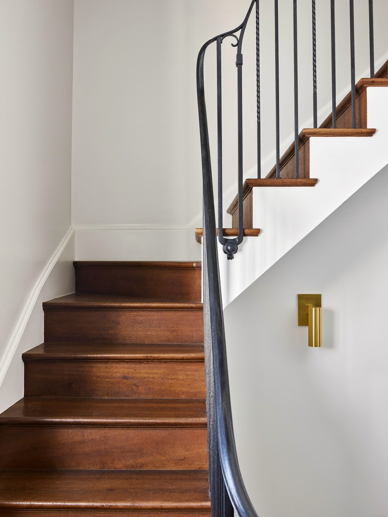 A renovated basement stair in an older Boston rowhouse: dark walnut treads, crisp white risers, simple black iron handrail on plain brackets, warm brass sconce at the turn, freshly painted walls