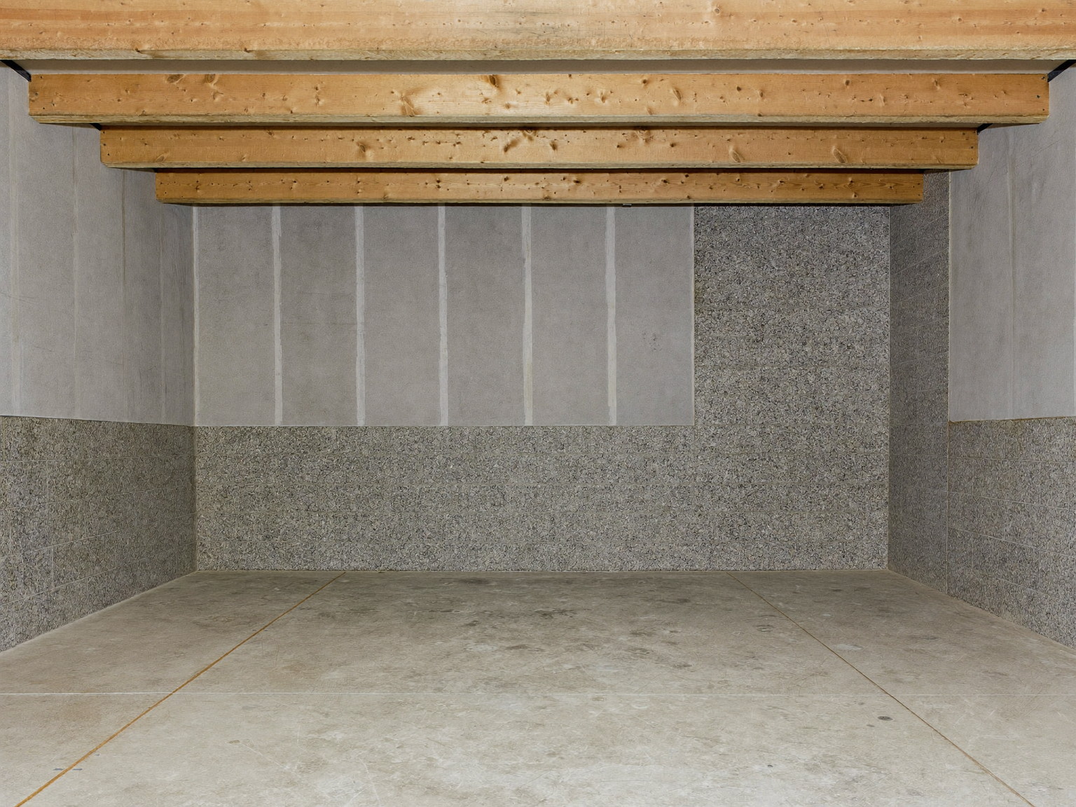 An unfinished rowhouse basement before renovation: low ceiling with exposed wood beams overhead, parged masonry foundation walls over a granite-flecked base, clean swept concrete floor, a single bare work light, completely empty