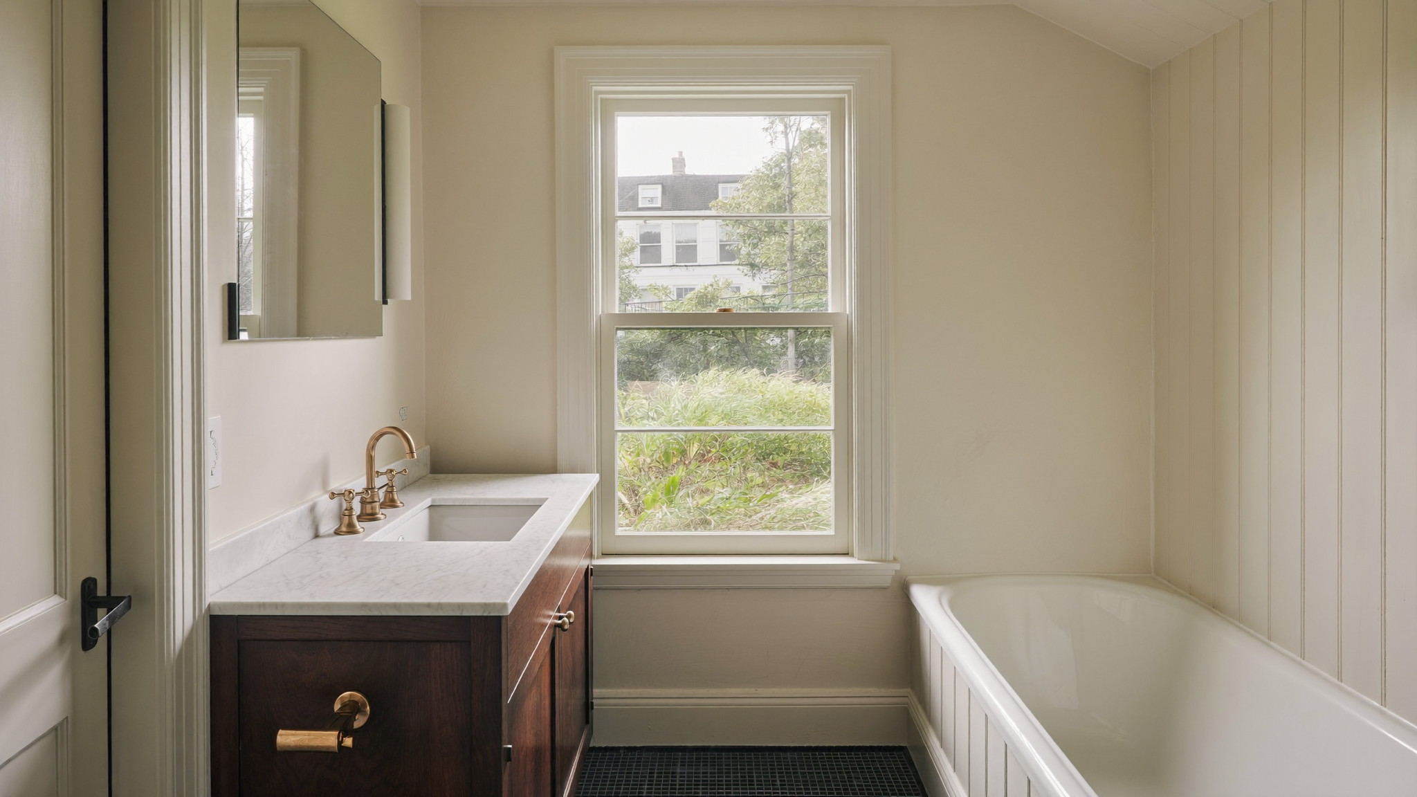 Editorial photograph of a renovated compact bathroom on an upper floor of a Federal-era Beacon Hill Boston rowhouse: ivory plaster walls, white marble-topped dark walnut vanity, unlacquered brass faucet, small white tub, tall six-over-six window with soft daylight