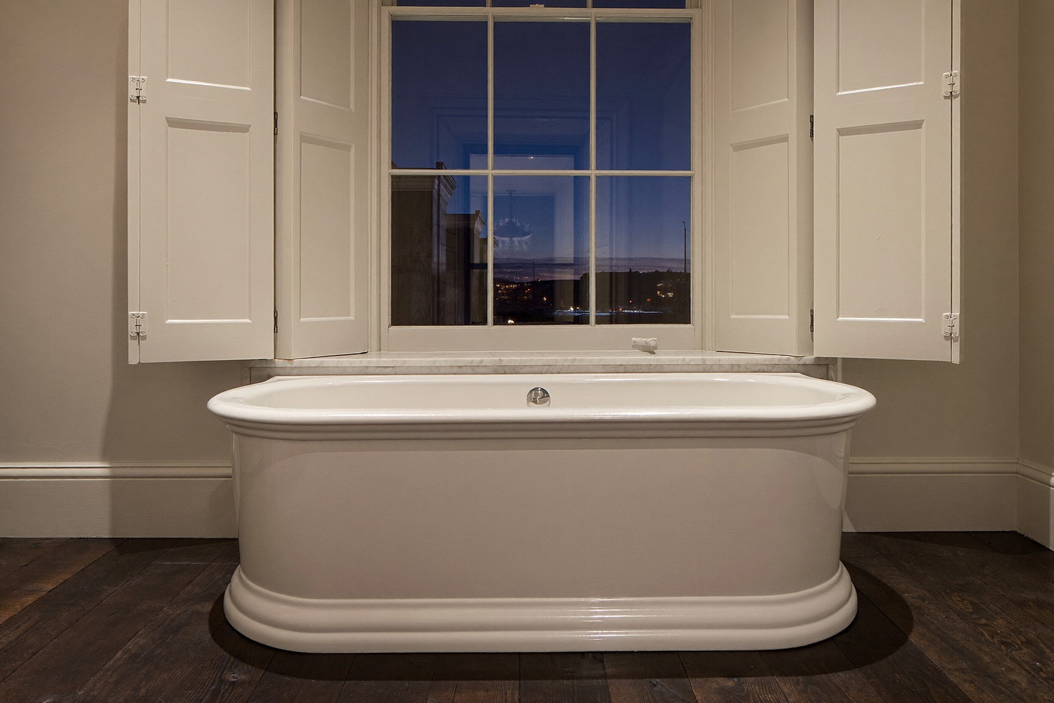 A deep white soaking tub beneath a tall shuttered window in a Beacon Hill Boston rowhouse bathroom, ivory walls, dark wide-plank wood floor, quiet evening lamplight, the tub rim completely bare
