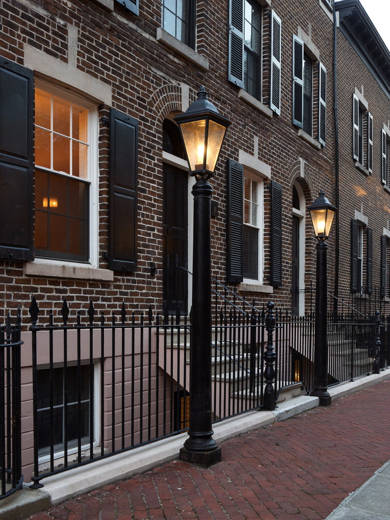 A gas-lantern lined brick sidewalk on Beacon Hill Boston in early evening: Federal-era brick rowhouses with black shutters and black iron handrails, warm windows beginning to glow, brick pavement