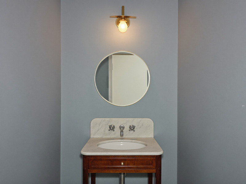 A compact powder room in an older Beacon Hill Boston rowhouse after renovation: soft gray-blue painted walls, small marble-topped walnut washstand, plain round brass mirror, brass sconce glowing warmly