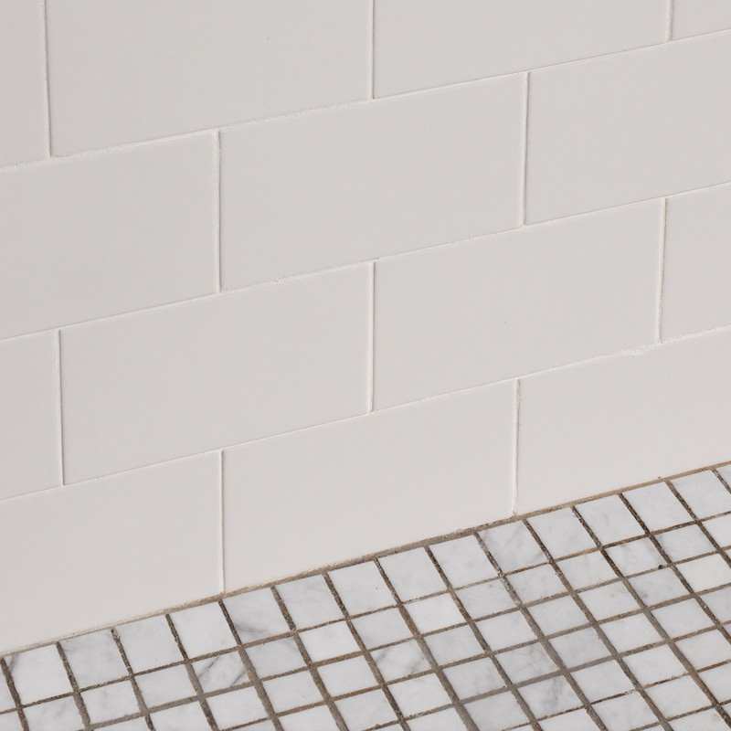Close view of classic white rectangular wall tile with crisp fresh grout lines meeting a strip of white carrara marble mosaic floor tile, precise straight joints, soft daylight