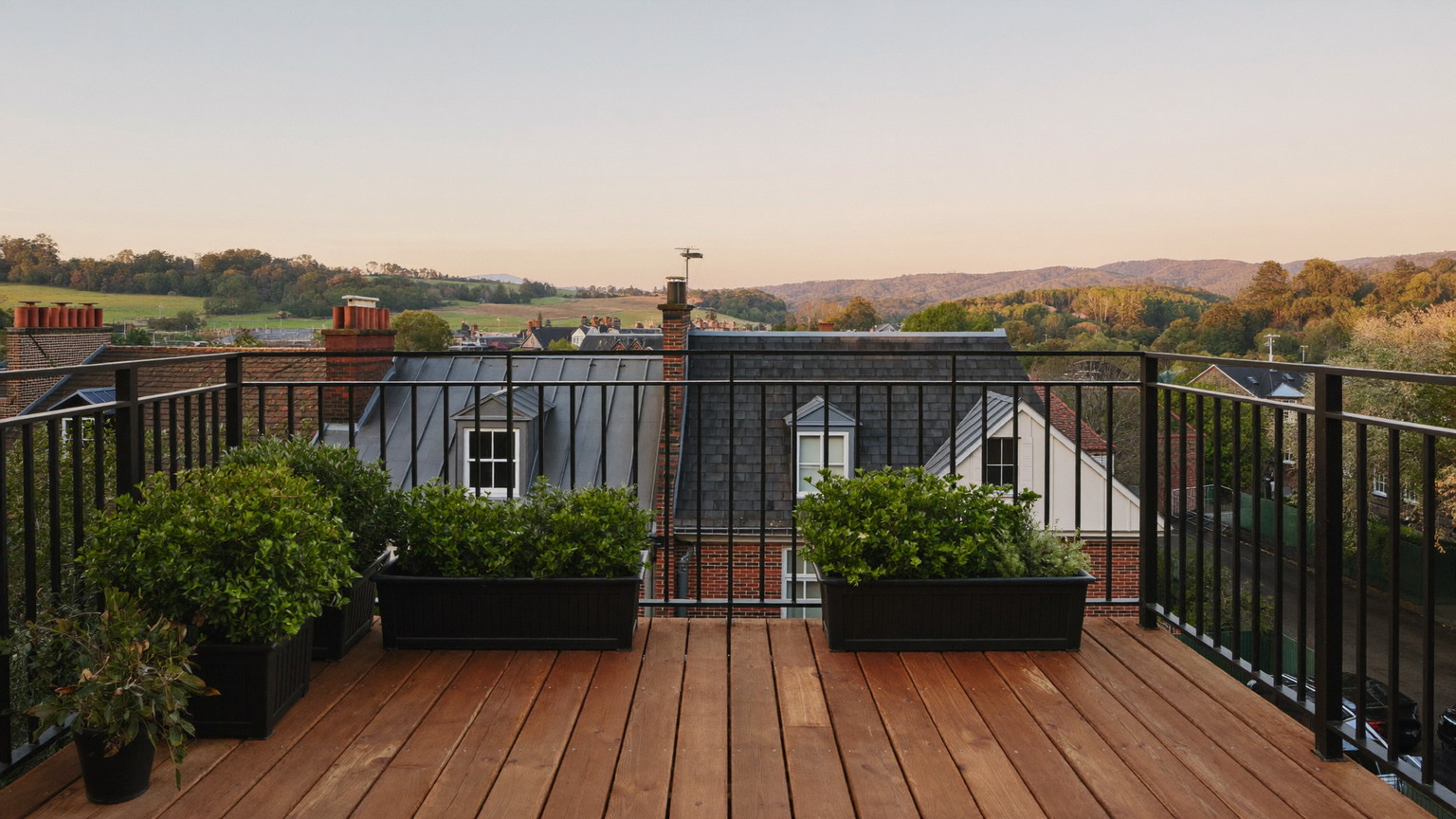 Editorial photograph of a compact rebuilt roof deck behind a Beacon Hill Boston rowhouse at golden hour: warm mahogany decking, black iron railing, potted boxwoods, brick chimneys and slate roofs beyond