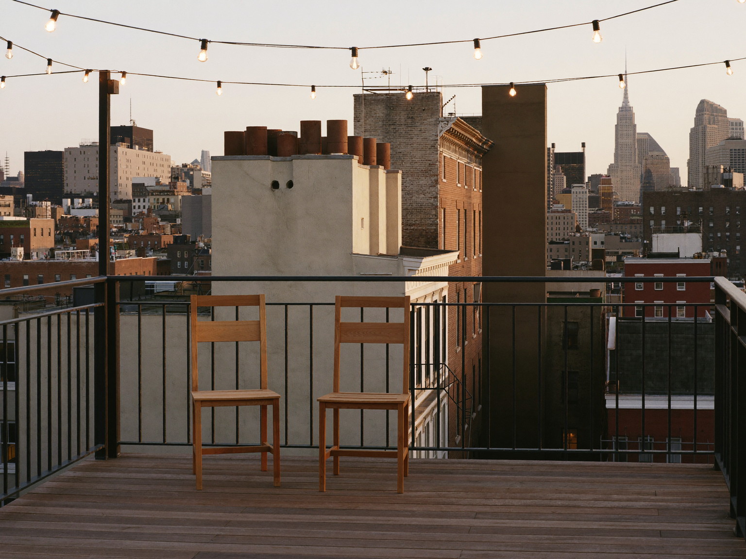 A finished compact city roof deck in warm evening light: oiled mahogany boards, black iron perimeter rail, two simple teak chairs facing the skyline chimneys, strings of plain warm bulbs overhead
