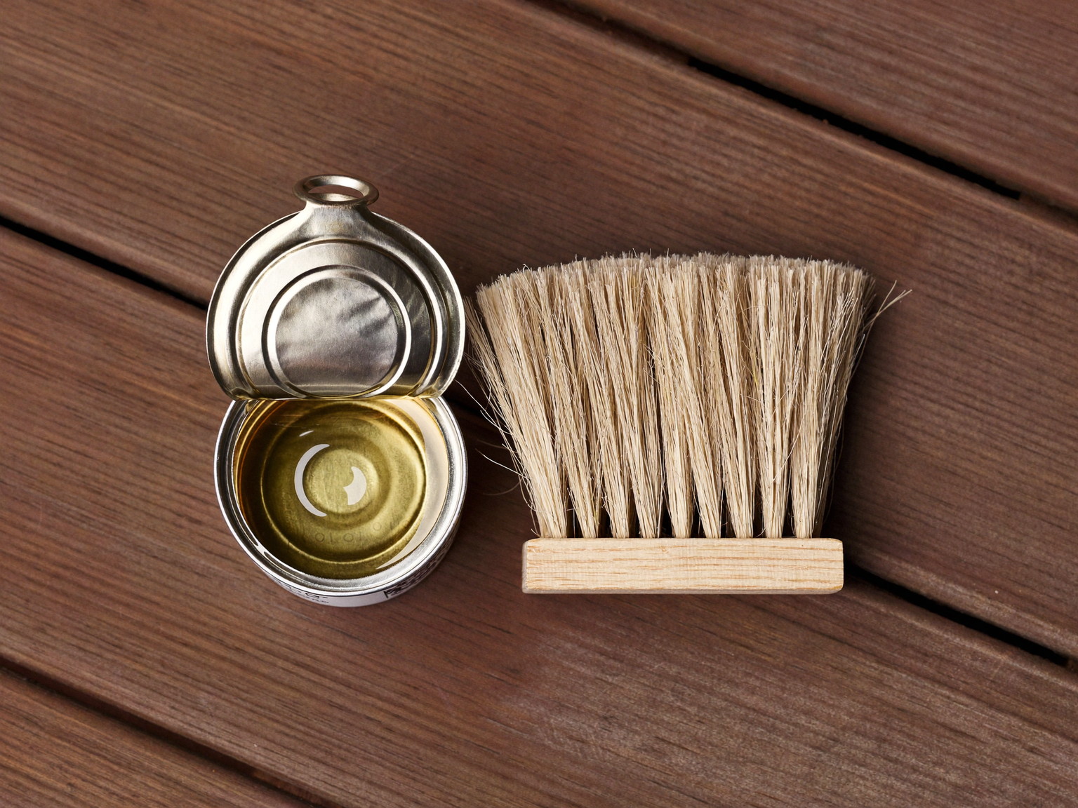 A hand applying nothing: a plain bare natural-bristle brush resting across an open unmarked metal can of clear penetrating oil on a mahogany deck board, seen from the side, completely unlabeled surfaces