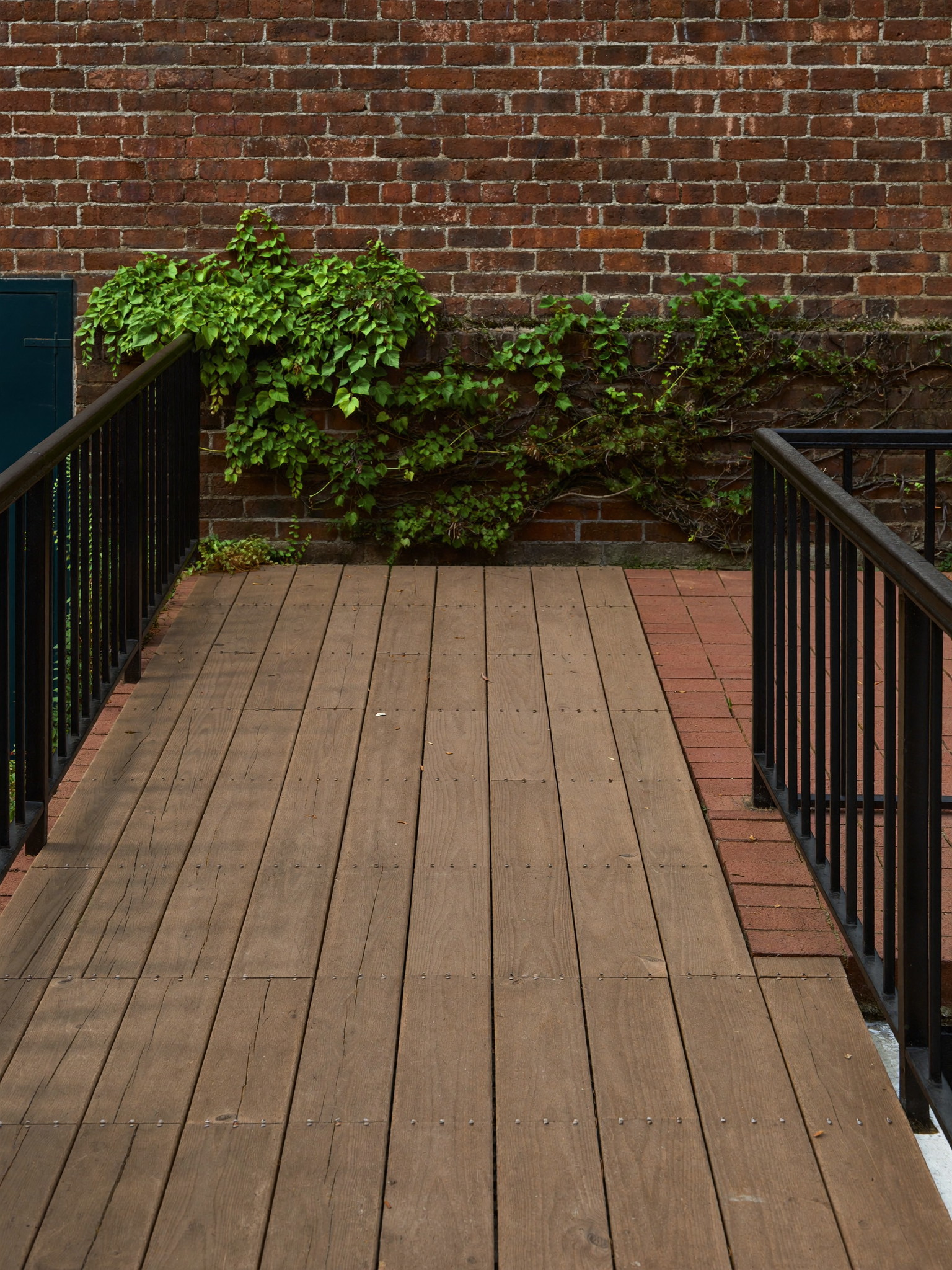 A small rebuilt rear terrace of a Boston rowhouse: new mahogany deck boards meeting old brick paving in a neat straight line, black iron rail, ivy on the brick garden wall, soft evening light