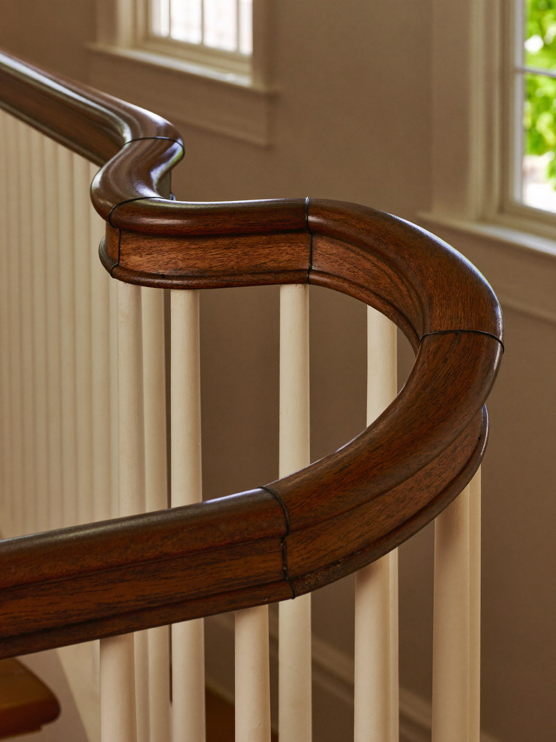 A repaired section of dark walnut stair handrail in an older Boston rowhouse, the new wood spliced and finished to match the original, white balusters below, warm window light