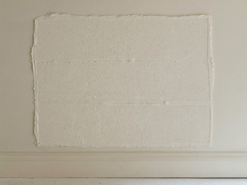 A smooth rectangular plaster patch on an ivory rowhouse wall, joint compound feathered wide and sanded flat, ready for paint, raking light showing the level surface, tidy floor below