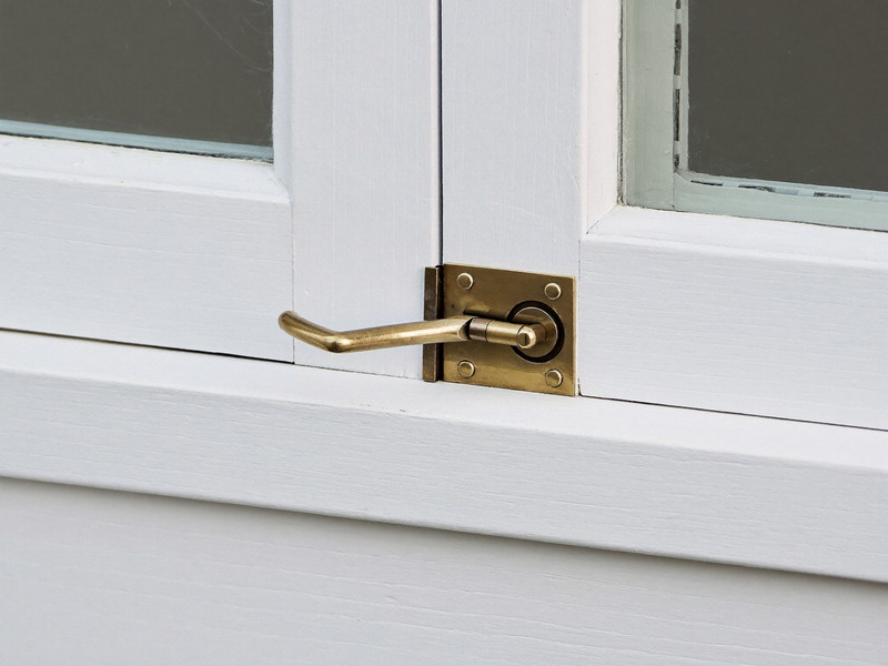 Close view of a traditional unlacquered brass window sash latch at the meeting rail of two white painted wooden window sashes, a small smooth sliding lever on a plain square base plate, crisp fresh white paint