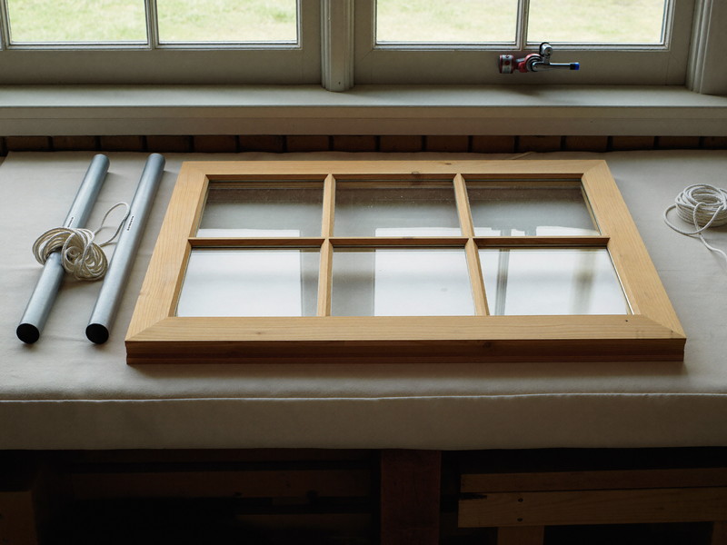 On a padded workbench in a rowhouse workshop, a wooden double-hung window sash lies flat, a neat loose coil of soft plain cotton cord and two plain smooth steel cylinders resting beside the sash