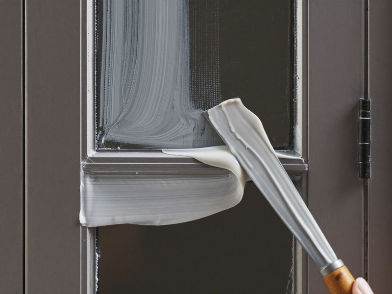 Close view of fresh glazing putty being tooled smooth along a single antique window pane, a plain putty knife mid-stroke, crisp clean glass, soft daylight
