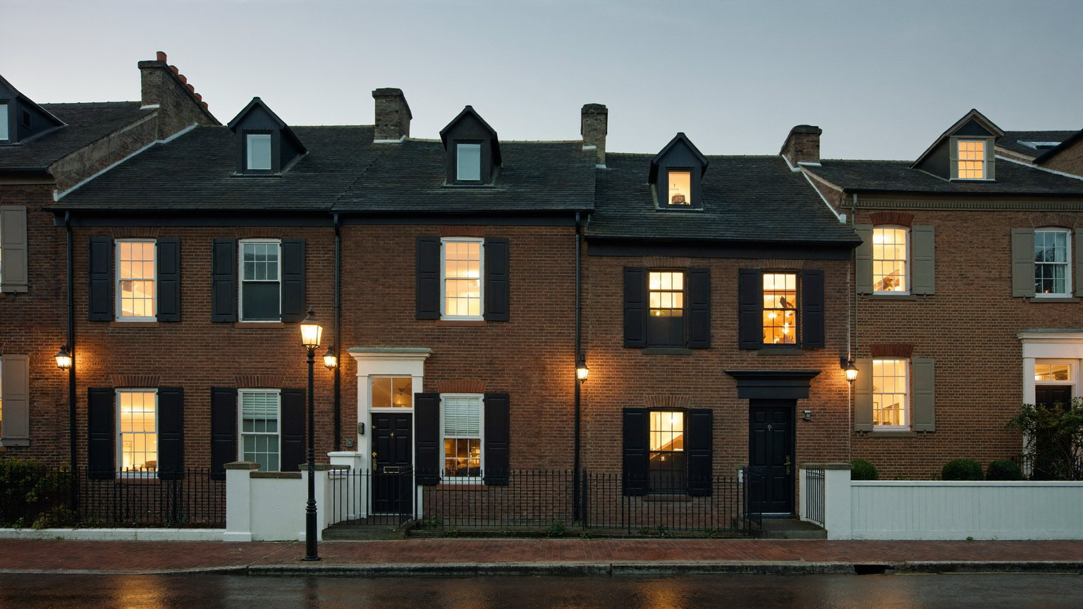 A quiet Beacon Hill Boston residential street in early evening: brick rowhouses with black shutters, gas lanterns glowing, brick sidewalk glistening after light rain, warm windows