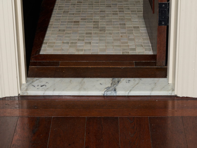 A small marble threshold freshly reset at a rowhouse bathroom doorway: white carrara marble strip sitting level between dark walnut floorboards and marble mosaic tile, crisp new grout lines