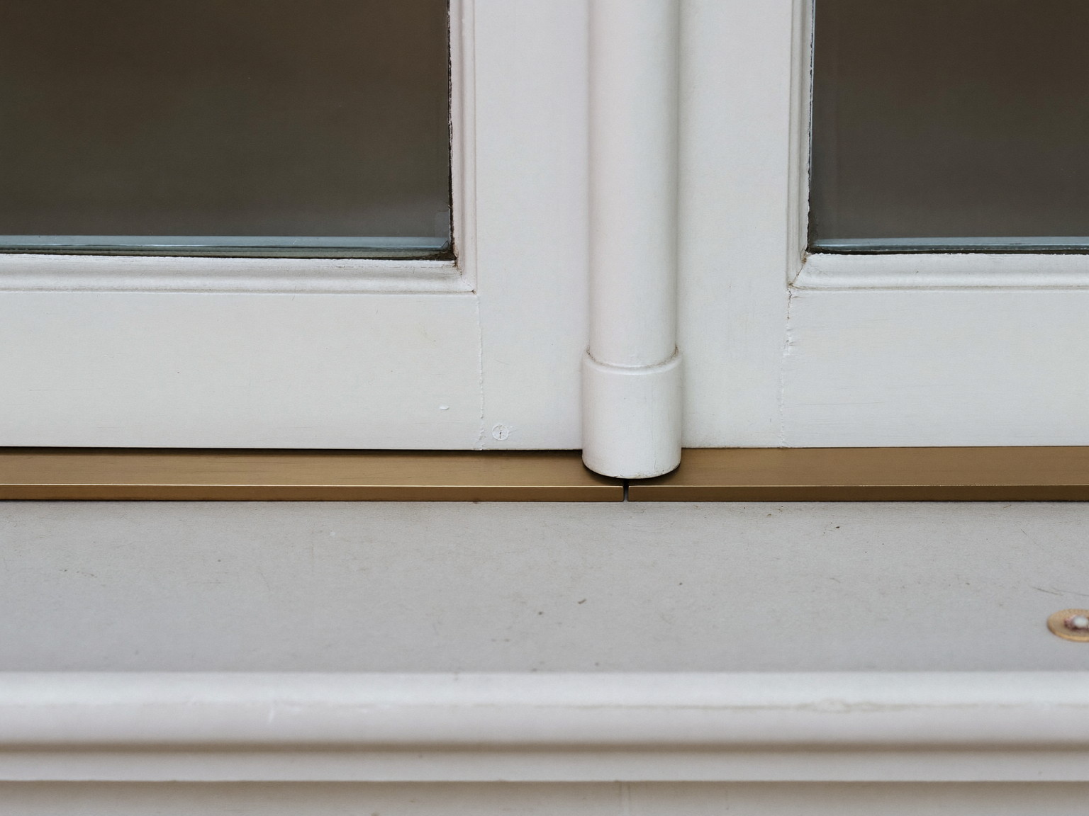 A slim bronze metal weatherstrip seated along the jamb and sill of an antique white painted double-hung window, the thin metal strip running straight and true, macro detail