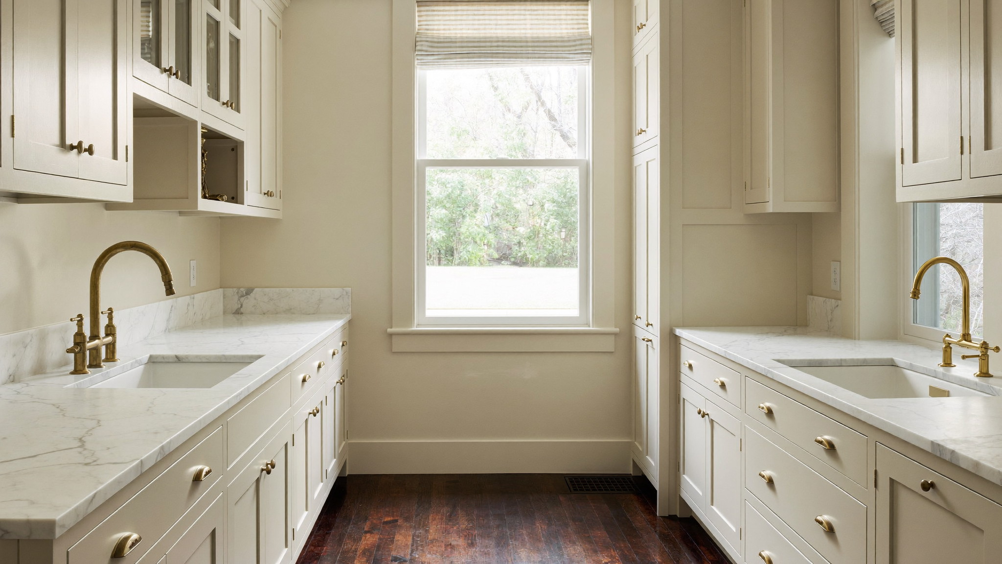 A renovated galley kitchen inside a Federal-era Beacon Hill Boston rowhouse, photographed editorially: ivory painted inset cabinetry, white carrara marble counters, unlacquered brass hardware, dark walnut floor, tall rear window with soft daylight