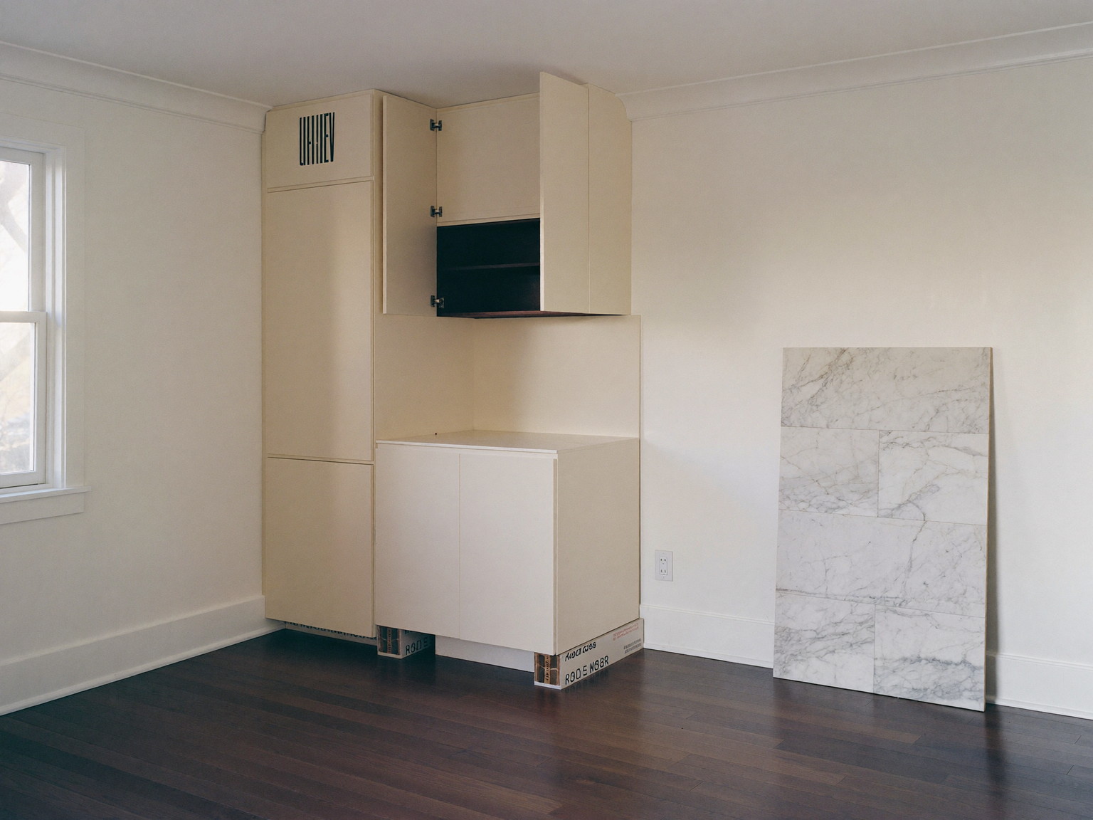 Mid-renovation compact rowhouse kitchen, tidy and completely empty with no one in it: new ivory cabinet boxes set level and clamped, marble slab template leaning against the wall on padded blocks, clean swept dark wood floor, soft daylight