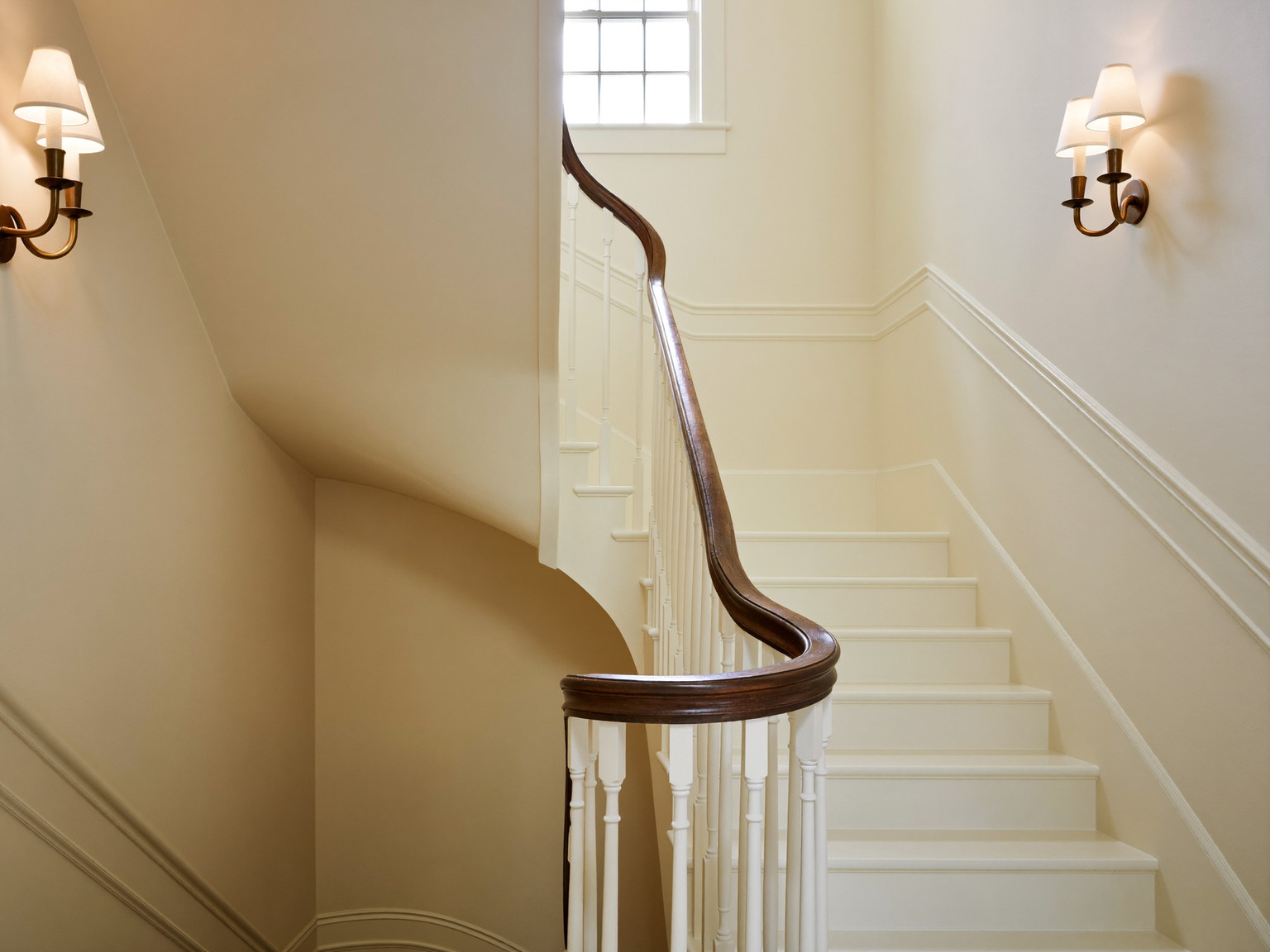 A repainted rowhouse stair hall: soft ivory walls, white balusters with a dark walnut handrail curving up the turn, restored wainscot, brass wall sconces, tall window light from above