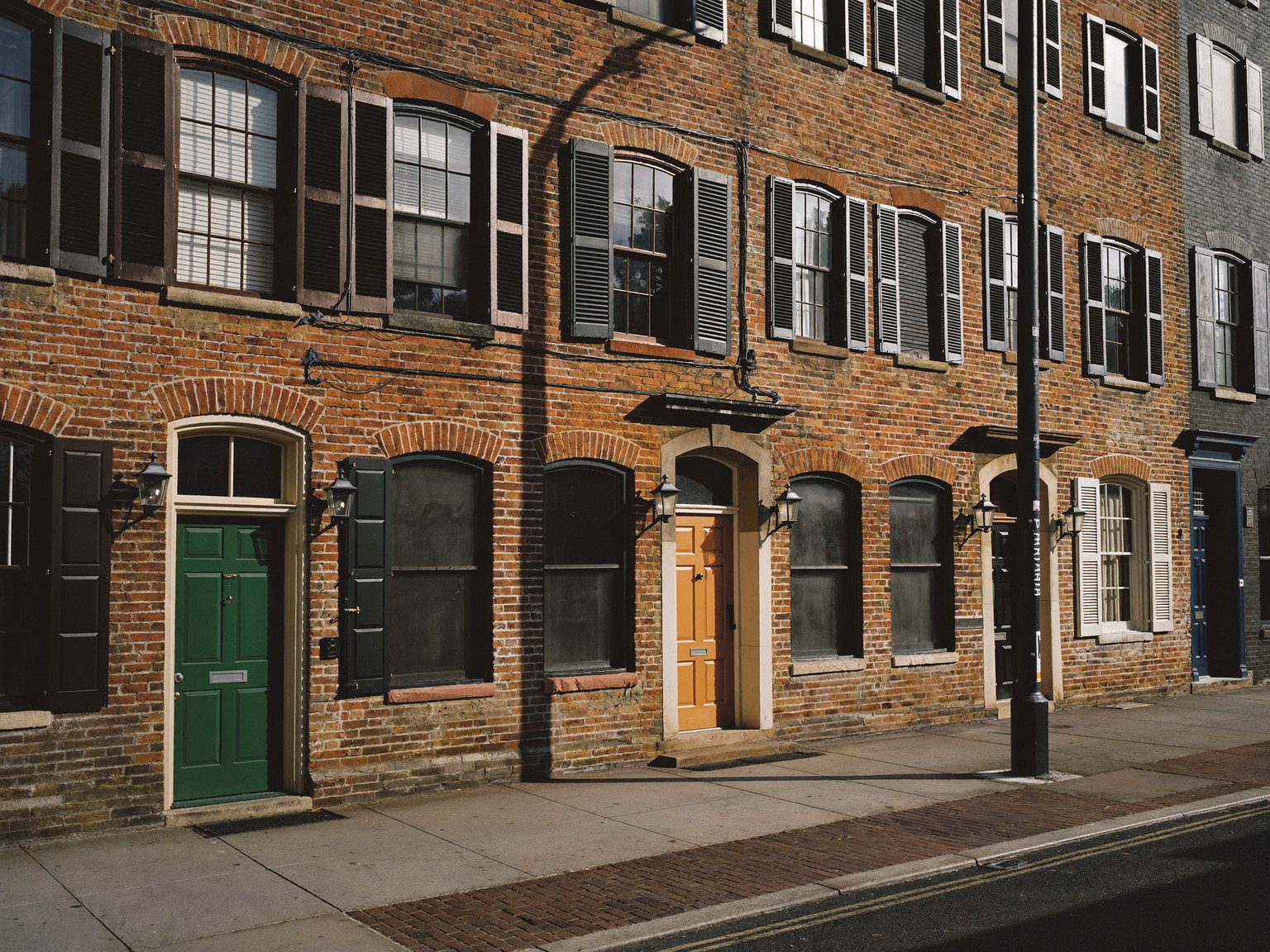 A row of Federal-era brick townhouses on a sloping Beacon Hill street: black shutters, deep-toned front doors in green, oxblood, and navy, gas lanterns, brick sidewalk, late afternoon light