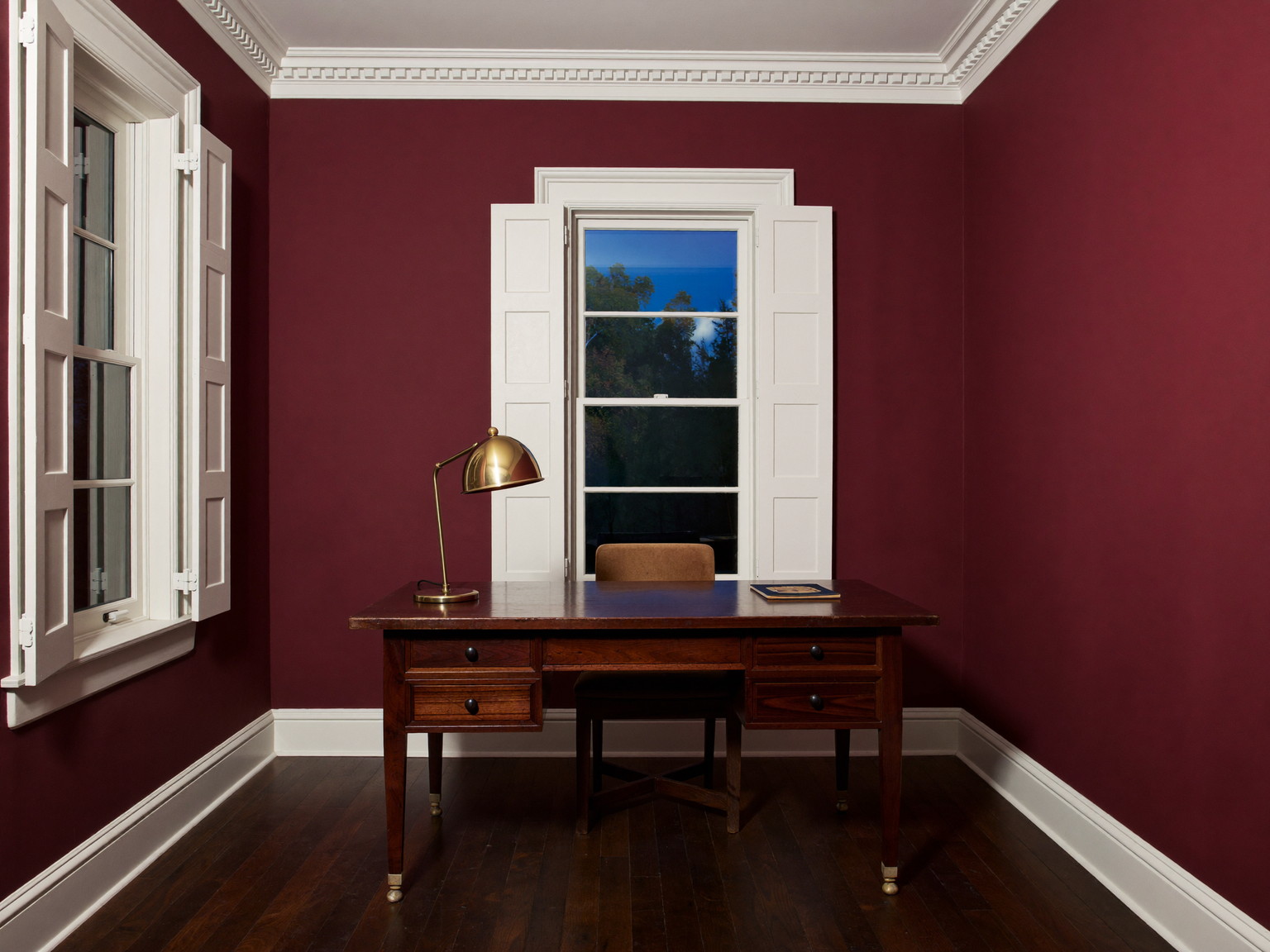 A freshly painted compact rowhouse study: deep oxblood walls with crisp white trim and restored shutters, dark walnut desk, brass lamp glowing, dark wood floor, evening mood