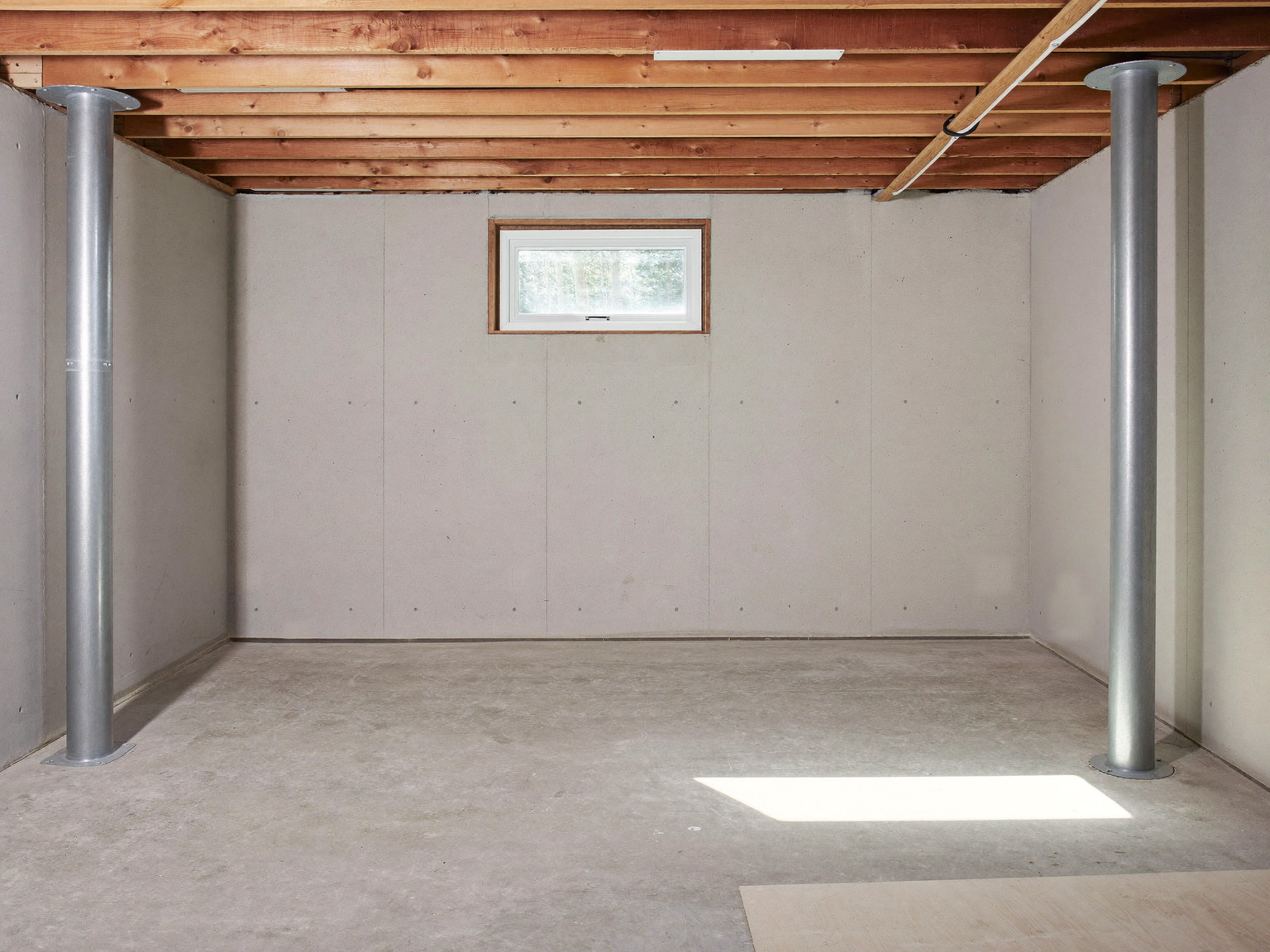 Clean unfinished New England basement with round lally columns, exposed joists, concrete walls, and a small hopper window