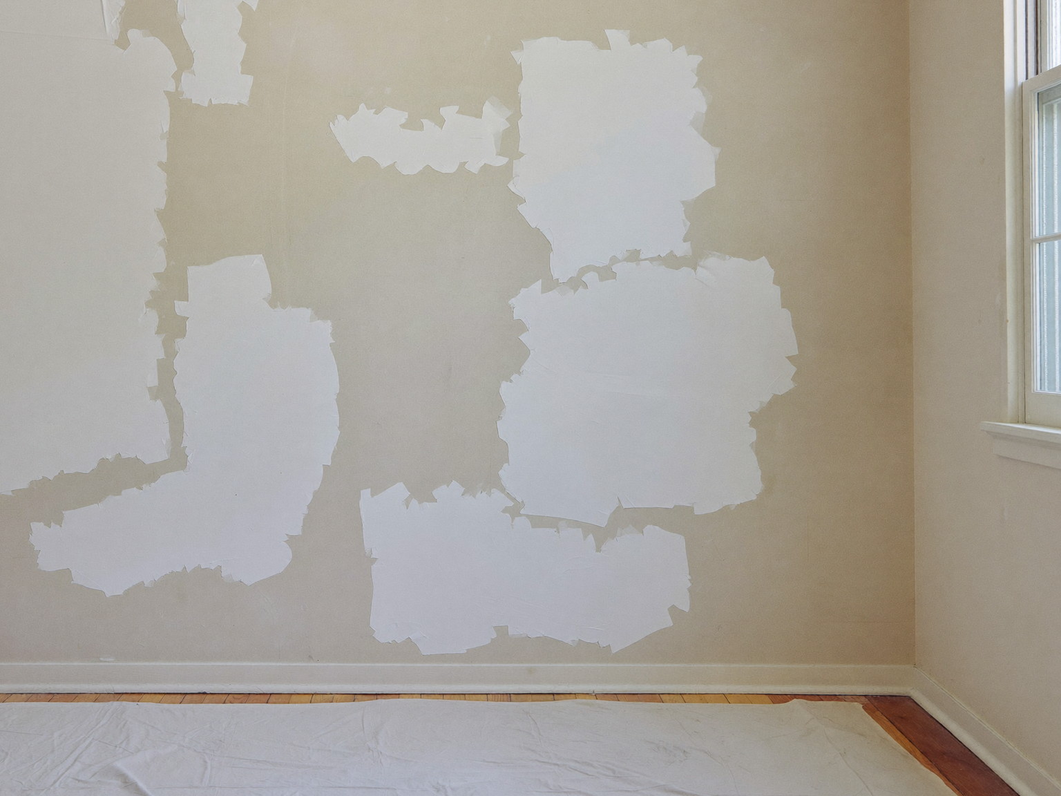 Older plaster wall mid-preparation: sanded joint compound patches and spot-primed repairs above a floor protected with a canvas drop cloth