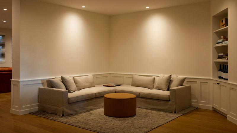 A warm finished lower-level den in an older city home: oatmeal linen sectional around a round camel leather ottoman