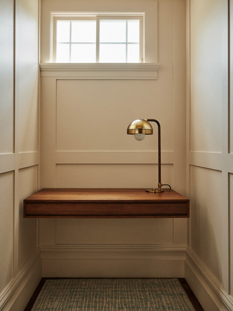 A compact lower-level study nook in an older home: warm putty-painted paneling, walnut floating desk with nothing on it, brass task sconce
