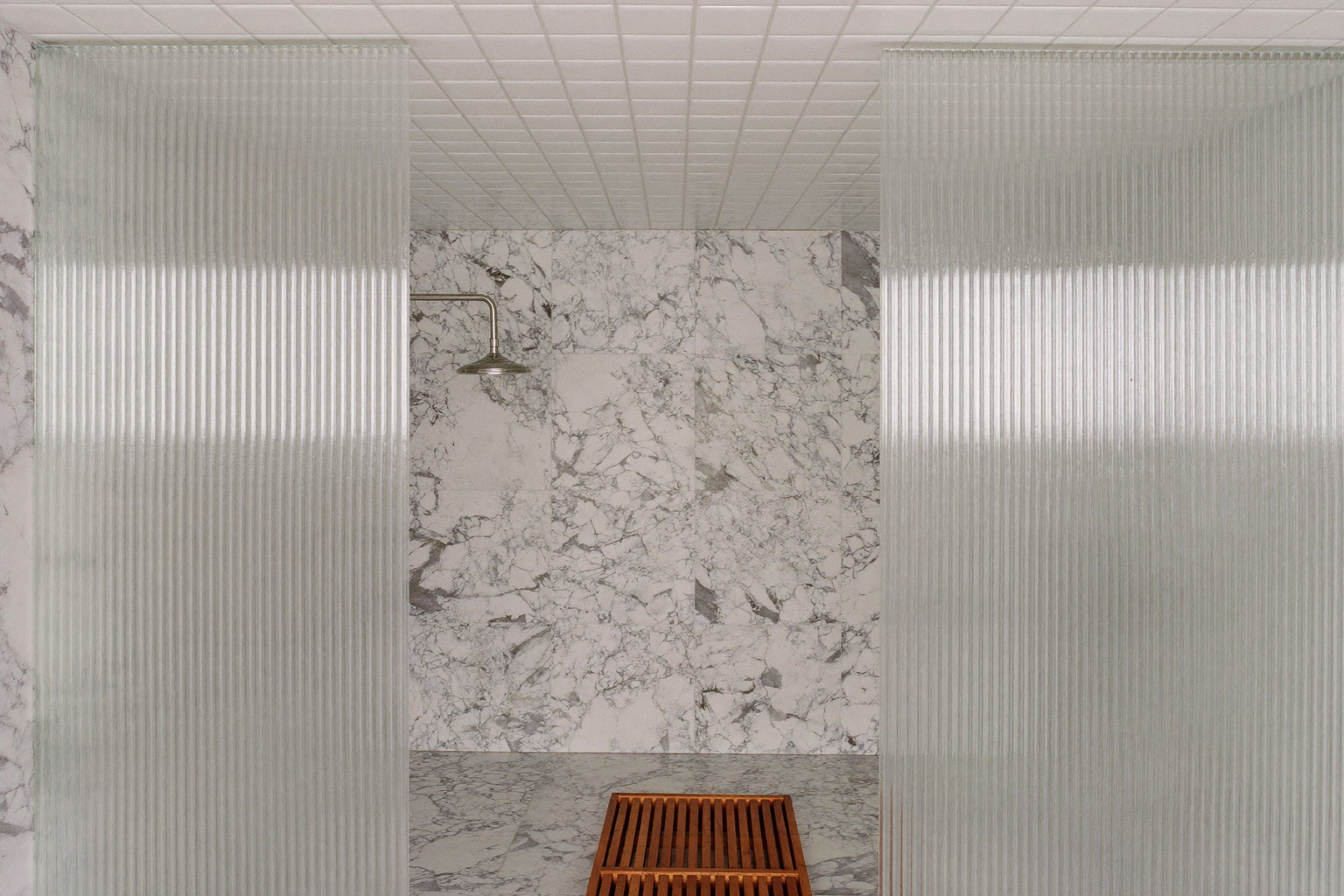 A compact marble wet room behind a fluted glass partition in a renovated older home, white ceramic square tile to the ceiling, polished chrome rain head
