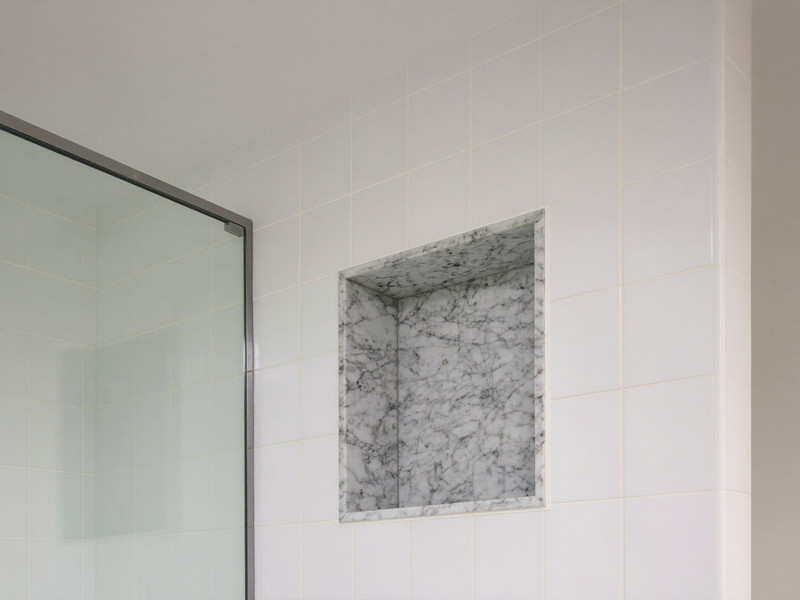 A walk-in shower detail: white ceramic tile to the ceiling, recessed niche lined in carrara marble with a slim chrome trim edge, spotless glass