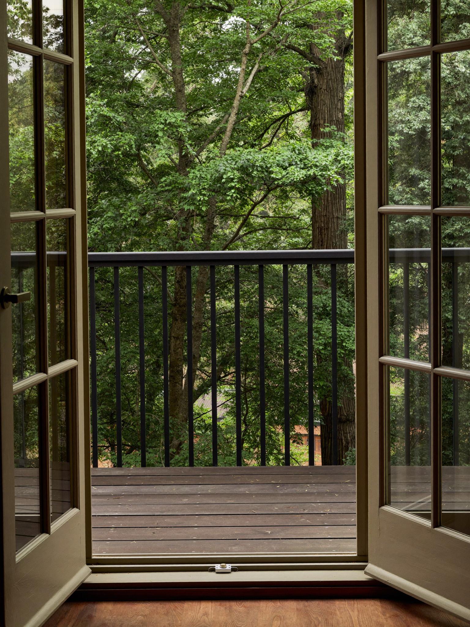 View through tall french doors from inside an older home onto a small cedar balcony deck with a dark charcoal metal railing, leafy mature trees beyond