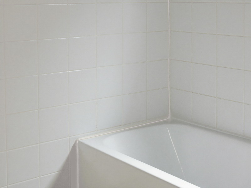 A clean even bead of fresh white caulk sealing the joint between a white tub edge and ceramic tile, tooled smooth, soft window light