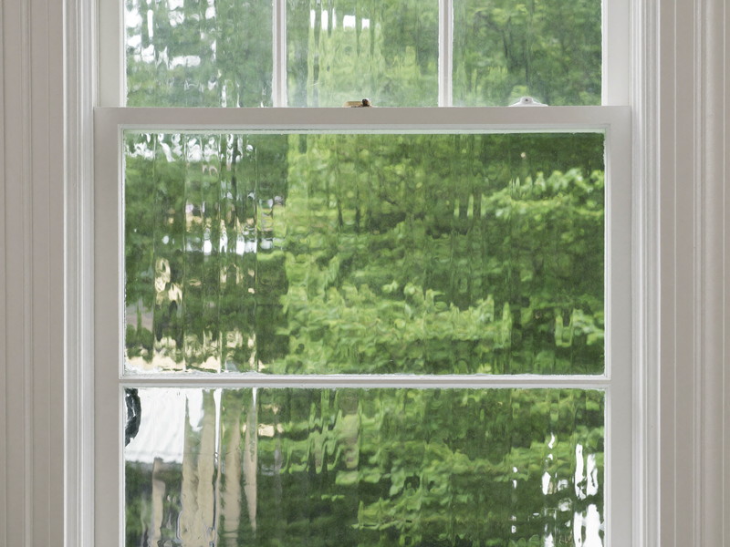 A restored double-hung window sash in an older home: fresh white paint, clean glazing, original wavy glass catching light, view of green leaves outside