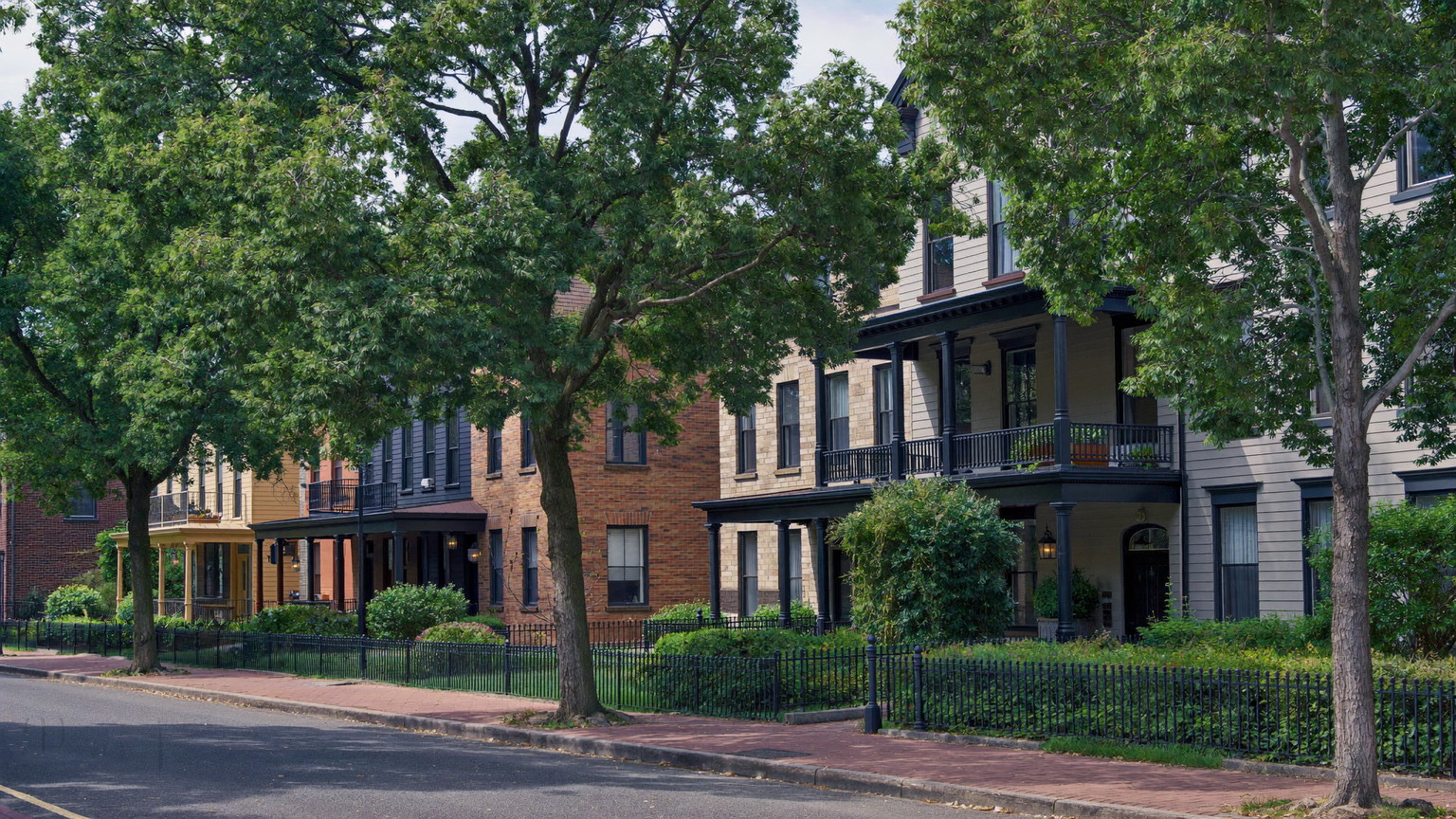 A quiet tree-lined Brookline-style street in summer: older brick and clapboard homes with generous porches, black iron fences, brick sidewalks, leafy canopy