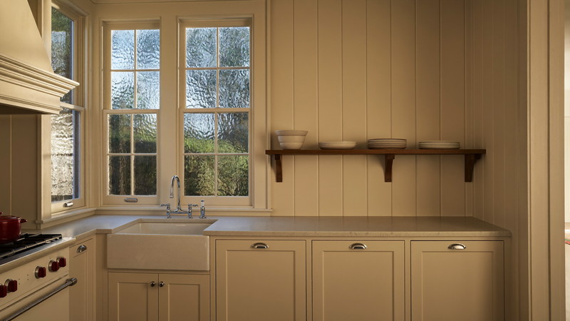 A renovated kitchen in an older Brookline-style home with original deep window casing and wavy glass, ivory cabinetry, walnut shelf with plain white dishes