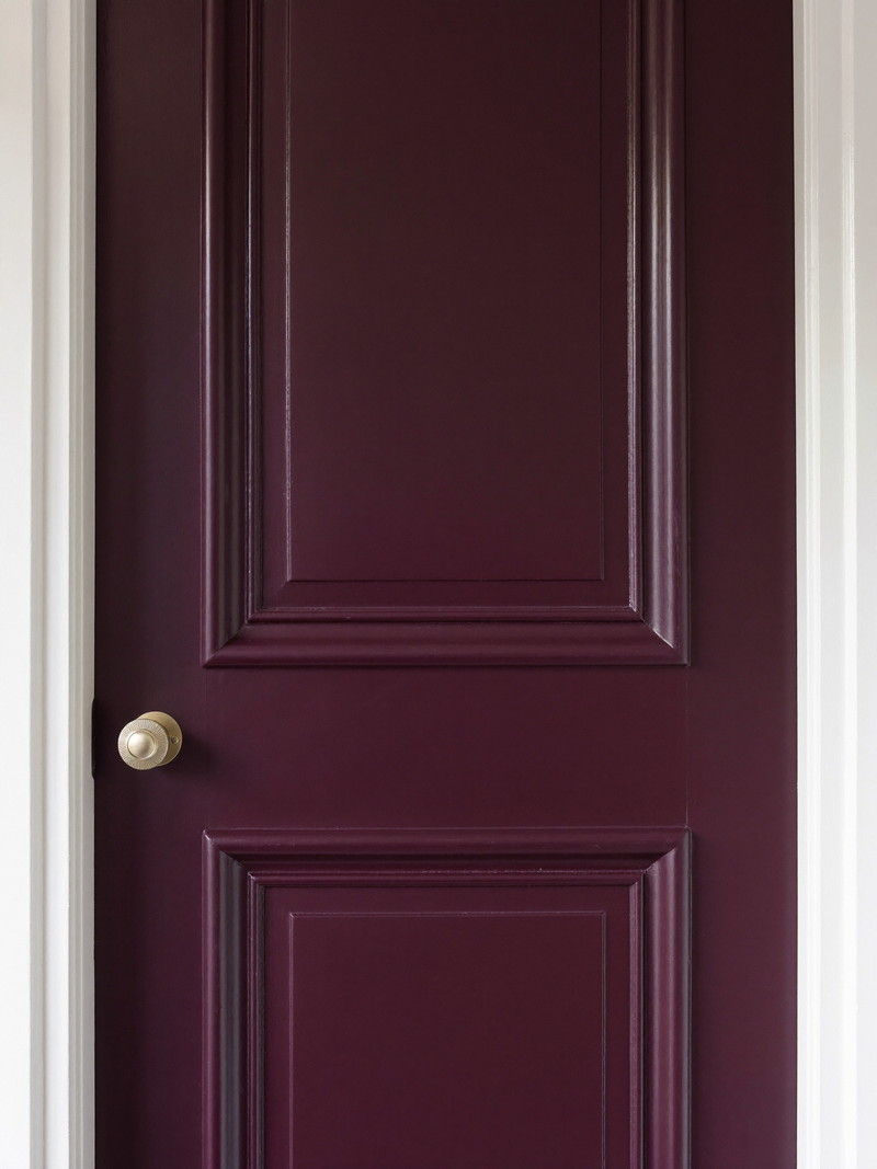 A paneled interior door freshly painted in deep plum-aubergine satin enamel with clean edges against restored white casing, plain unmarked brass knob