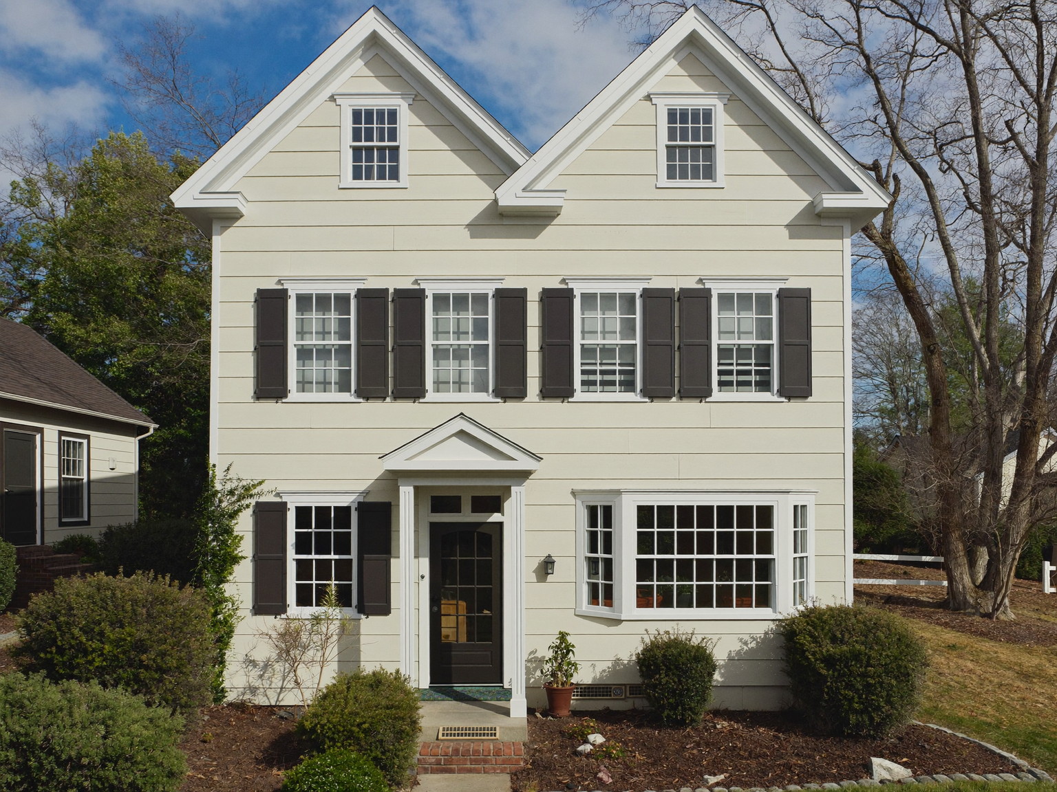 An older Brookline-style home exterior after a complete repaint: warm ivory clapboards, white trim, dark charcoal shutters, mature landscaping