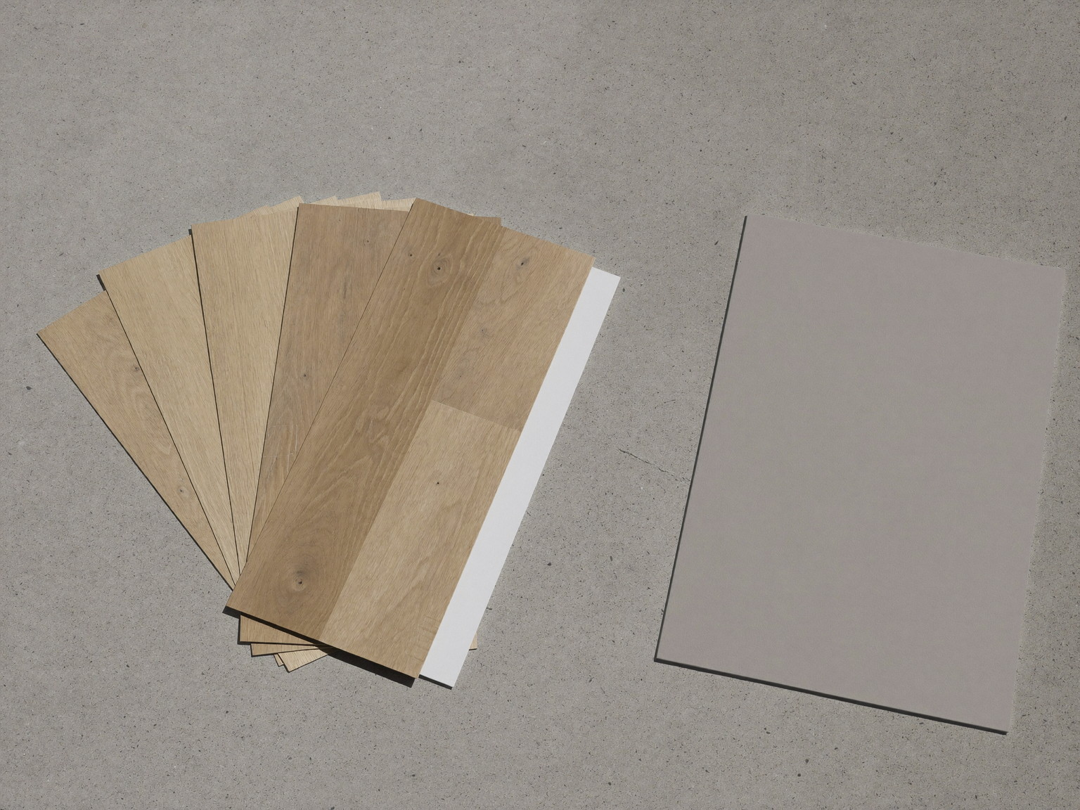 Overhead photograph of oak-look plank flooring samples fanned out on clean bare concrete beside a plain gray insulation board with a bare smooth face, soft
