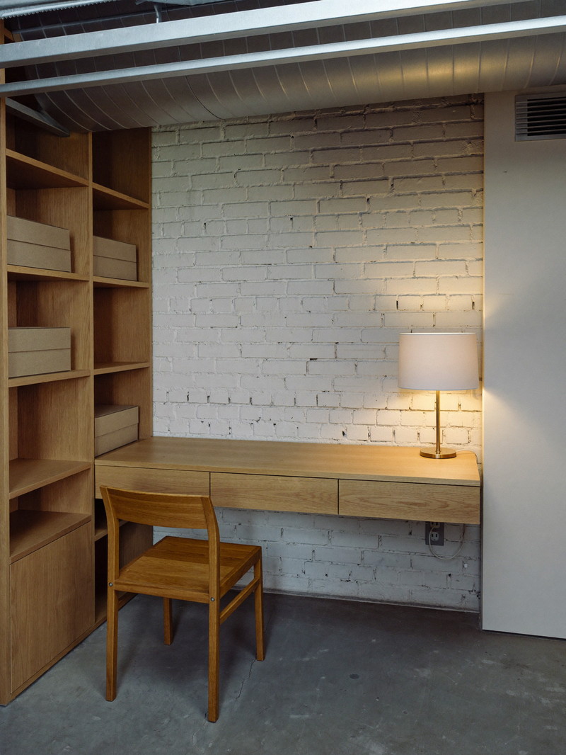 A compact lower-level home office nook in a renovated urban basement: built-in white oak desk against a warm white painted brick wall, simple wooden chair