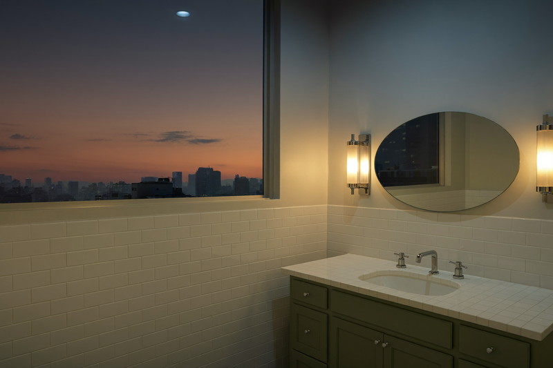 A calm finished city bathroom at dusk: chrome sconces lit beside an oval mirror, sage-green vanity, white carrara top, white stacked-bond tile glowing warm