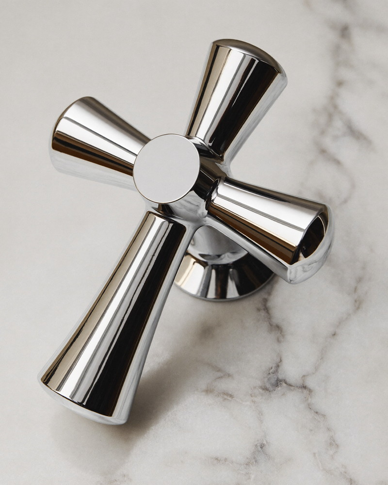 Macro photograph of a polished chrome cross-handle tap mounted on white carrara marble, smooth unengraved metal faces, photographed at a slight angle with soft