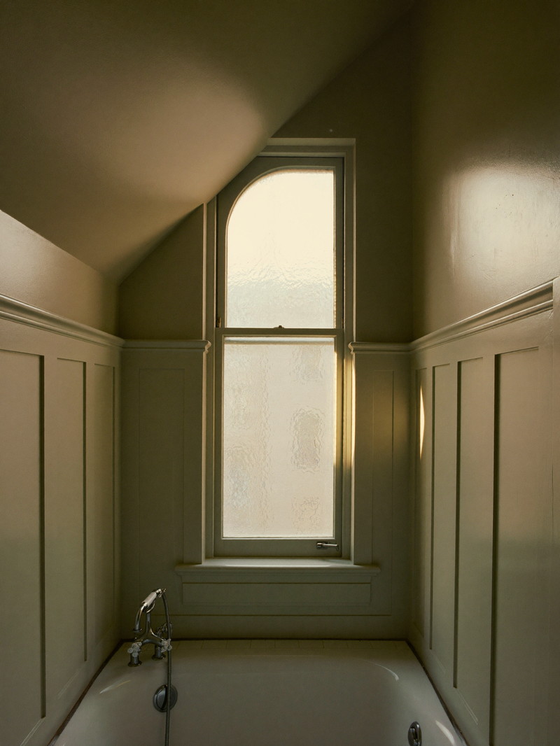 A renovated narrow bathroom on the top floor of an older Cambridge triple-decker: sloped ceiling over a white tub, sage-green paneling, tall narrow window with