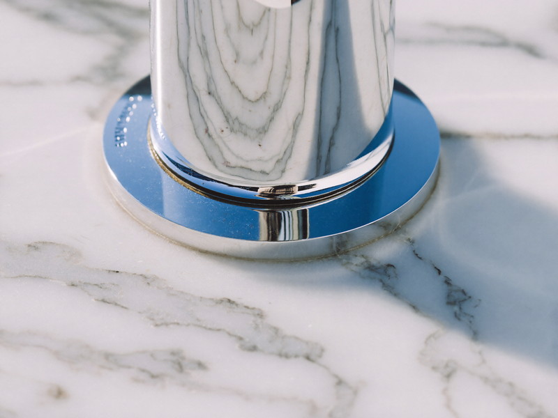 Macro photograph of white carrara marble with soft gray veining meeting a polished chrome smooth-faced faucet base, shallow depth of field, bright natural light
