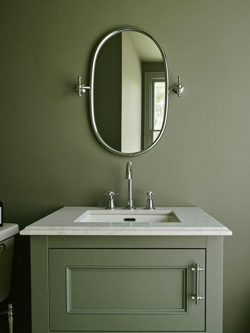 A sage-green painted bathroom vanity with white carrara marble top and polished chrome faucet with smooth cross handles, oval chrome-framed mirror on a smooth