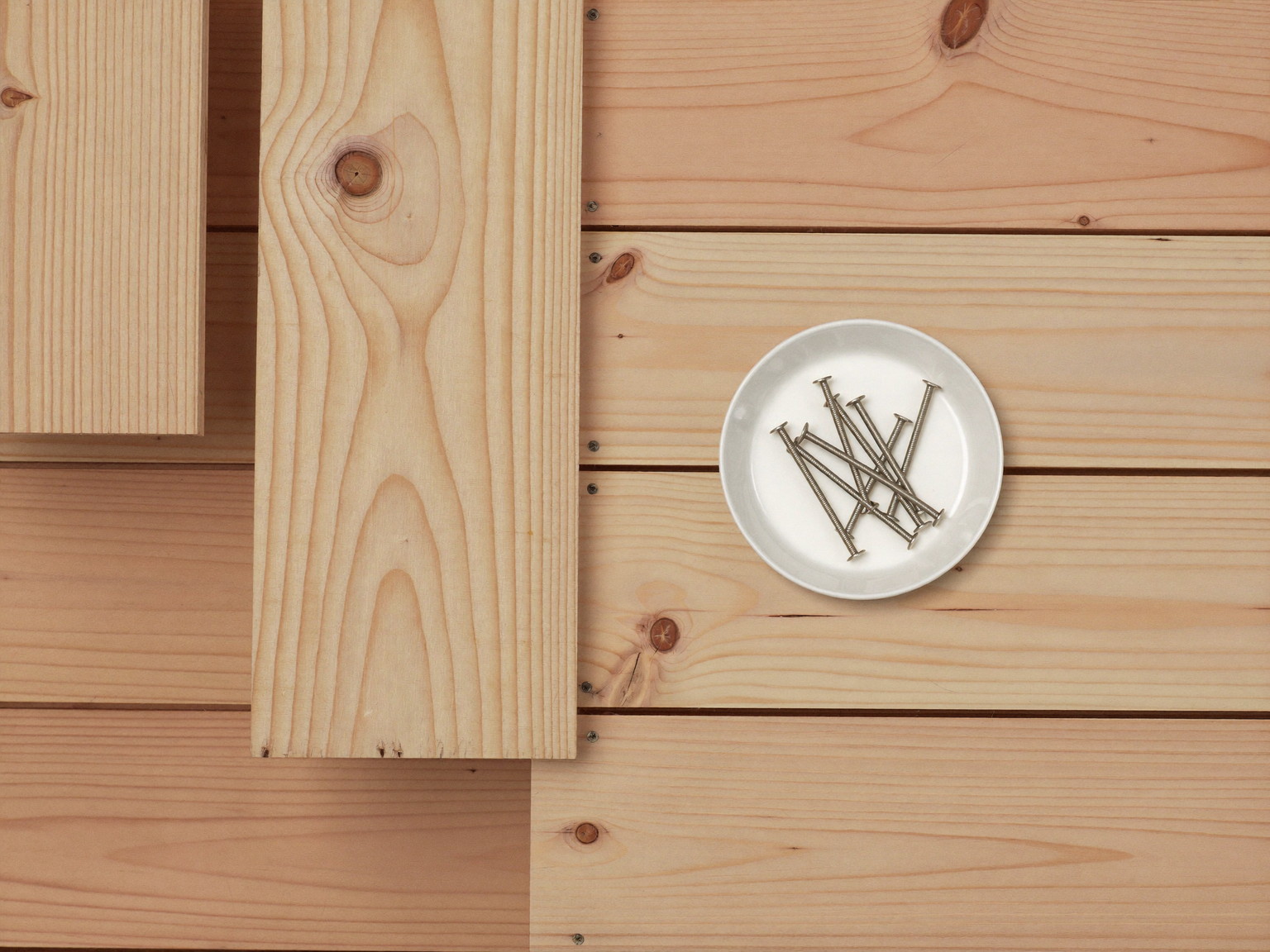 Overhead macro of new clear fir decking boards stacked neatly, smooth bare wood grain, a small plain white ceramic dish of stainless steel finish nails beside
