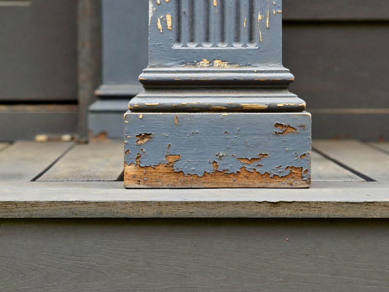 Close-up of an aged porch post base on an older city home before repair: layers of peeling gray paint, soft weathered wood grain at the bottom, honest wear