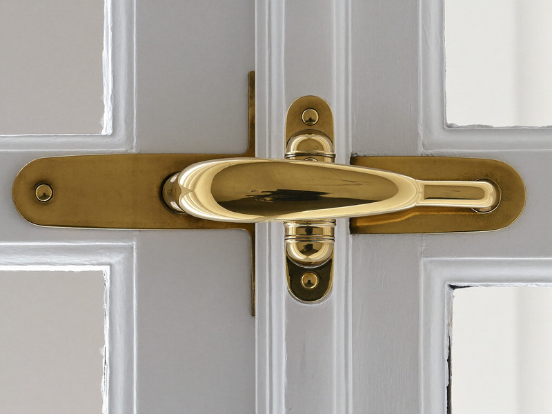 A traditional brass window sash lock with a curved swing arm mounted where two white painted wooden window sashes meet, seen close up, smooth polished brass