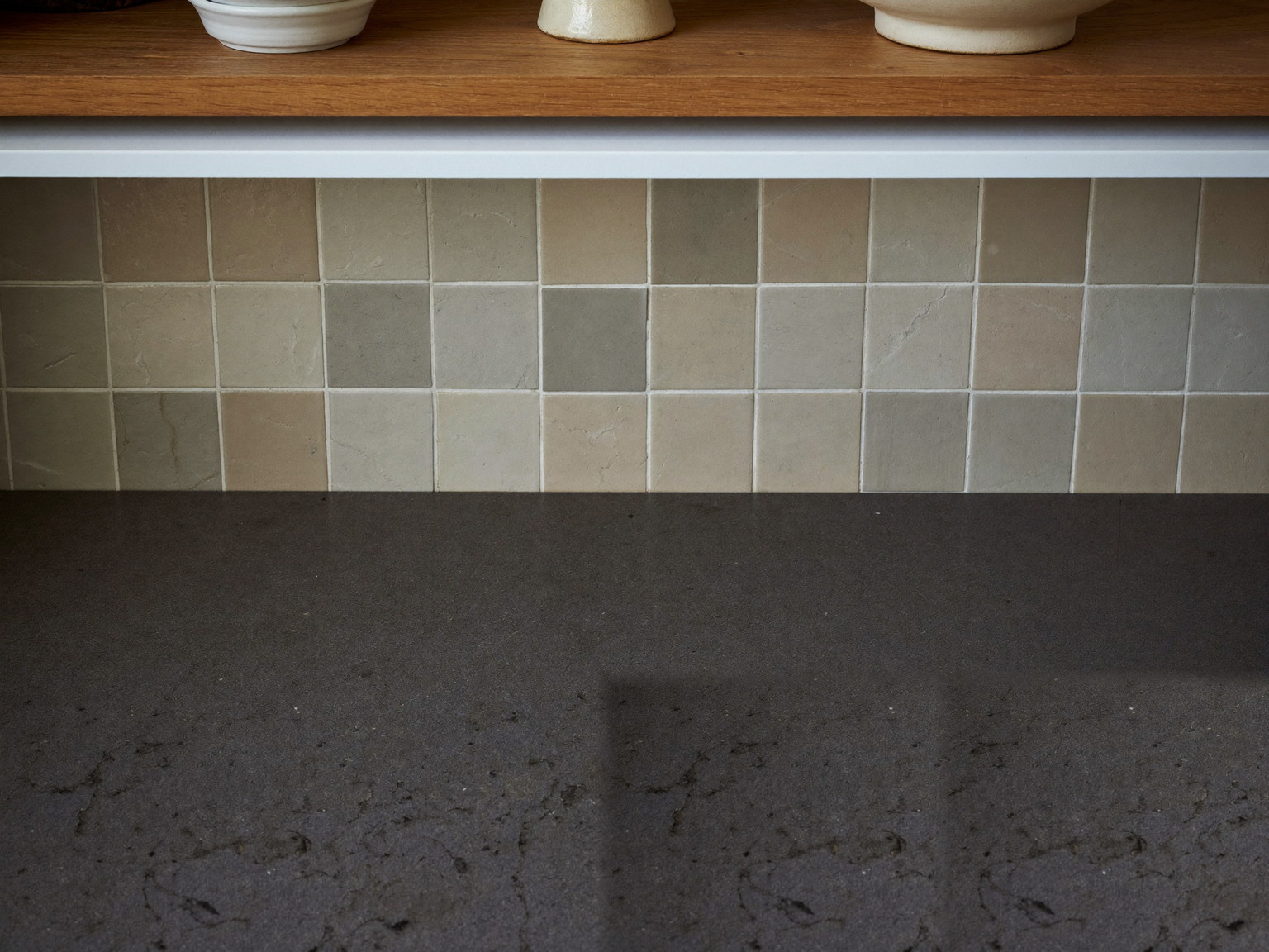 Macro photograph of a charcoal soapstone counter with an oiled sheen meeting a cream square handmade tile backsplash, a slim white oak shelf above holding